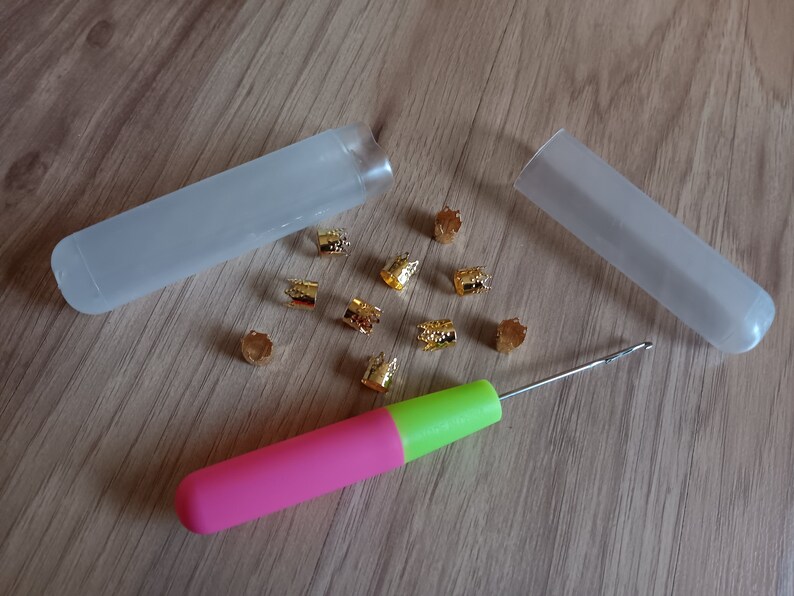 Może przedstawiać: Zestaw złotych koralik&oacute;w do włos&oacute;w w dw&oacute;ch przezroczystych plastikowych pojemnikach, wraz z r&oacute;żowym i zielonym haczykiem do szydełka.