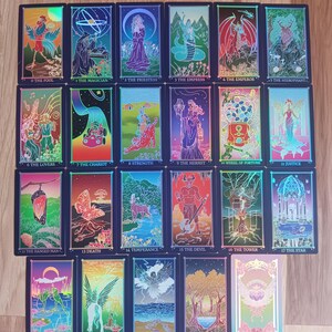 May include: A collection of 22 tarot cards, each featuring unique illustrations and titles. The cards have a dark blue border with vibrant artwork depicting figures, symbols, and scenes. The titles include "The Fool," "The Magician," and "The Lovers."