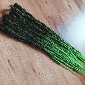 Könnte beinhalten: Ein Satz synthetischer Dreadlocks mit einem Ombre-Effekt von Schwarz zu Grün. Die Dreadlocks sind lang und gerade.