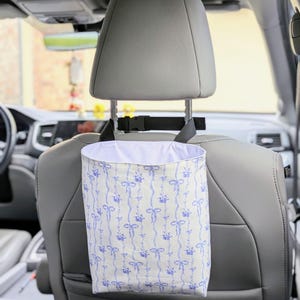 May include: A white car trash bag with a blue floral and bow pattern hangs from a car headrest. The bag is made of fabric and has a white interior. The car seat is gray.