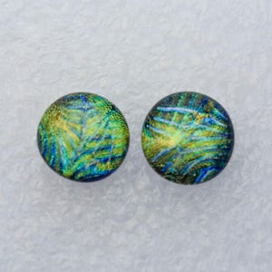 May include: Two round dichroic glass stud earrings. The earrings feature a vibrant mix of colors, including blue, green, and gold, creating a shimmering, iridescent effect. The earrings are set against a white background.