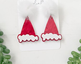 May include: Red Santa hat earrings with white fluffy pom-poms and white trim. The earrings are displayed on a white card, perfect for holiday or Christmas themed jewelry.