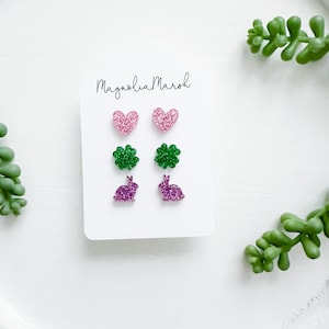 May include: A set of six glitter earrings on a white card with the text "Magnolia Maroh". The earrings are in the shapes of hearts, four-leaf clovers, and bunnies. The hearts are pink, the clovers are green, and the bunnies are purple.