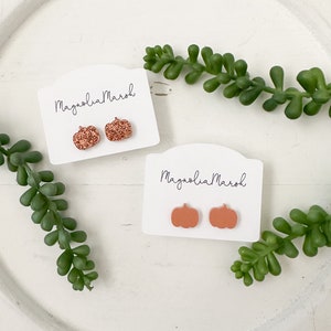 May include: Two pairs of pumpkin-shaped earrings on white cards. One pair is a glittery copper color and the other pair is a solid orange color. The cards have the text "Magnolia Marsh" written on them.