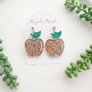 May include: A pair of apple-shaped earrings with a glittery, colorful design. Each earring features a green leaf and gold-colored post. The earrings are displayed on a white surface with the text "Magnolia Marsh" on the product tag.