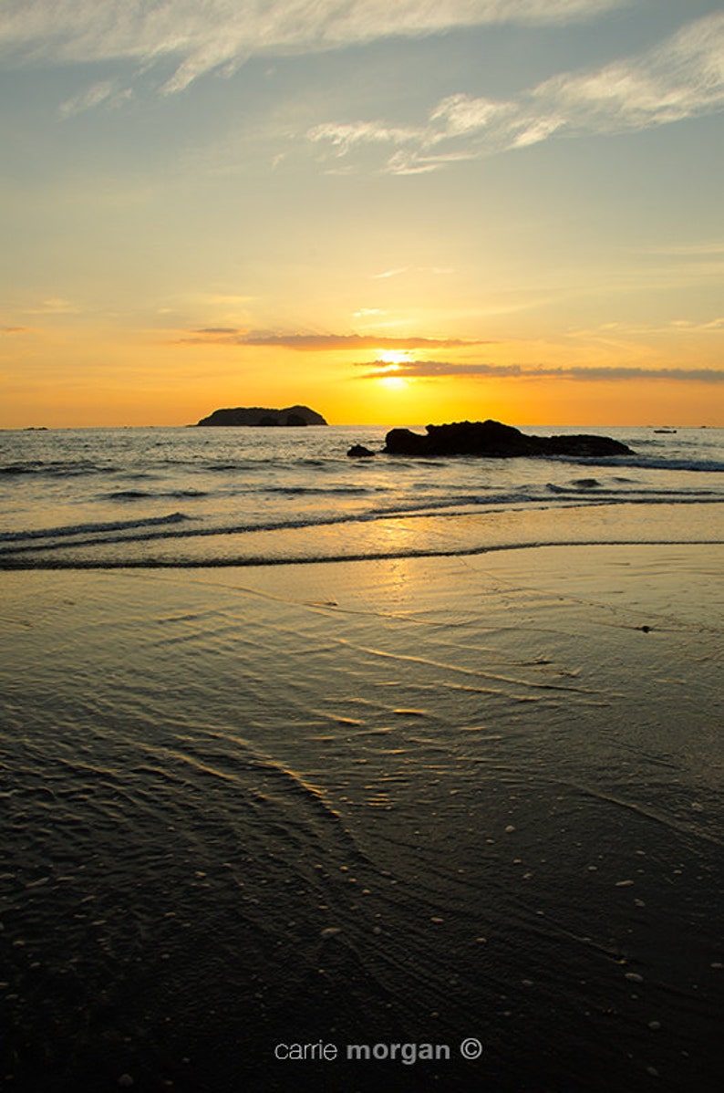 May include: A scenic sunset over a calm ocean with a small island in the distance. The sky is a vibrant orange and yellow, with soft clouds. The water is a deep blue, reflecting the colors of the sky. The beach is sandy and wet, with ripples from the receding tide.
