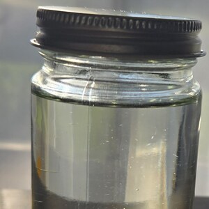 May include: A clear glass jar with a dark metal lid, filled with a transparent liquid. The jar is cylindrical with a slightly textured surface. The liquid appears to be clear and clean. The jar is sitting on a dark surface.