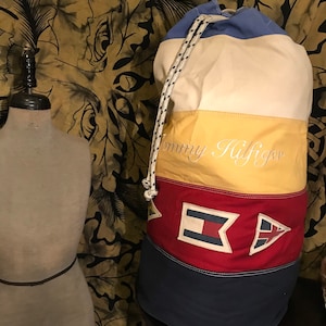 Puede incluir: Una bolsa de lona grande, blanca, azul, roja y amarilla con cierre de cordón. La bolsa tiene el texto "Tommy Hilfiger" bordado en la parte delantera. La bolsa tiene un tema náutico con banderas rojas, blancas y azules en la parte delantera.