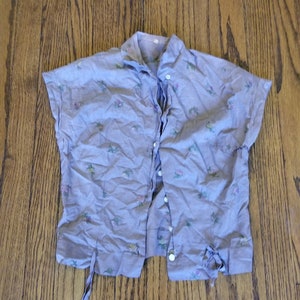 May include: A light purple short-sleeved button-up shirt with a floral print. The shirt has a tie at the waist and a button-up front.