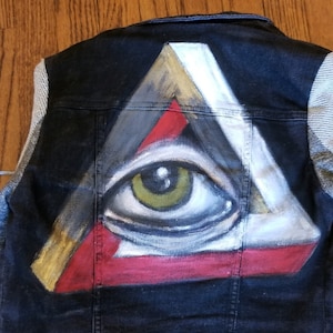 May include: A black denim jacket with grey sleeves and a painted all-seeing eye design on the back. The eye is inside a triangle with gold, red, white, and grey accents.