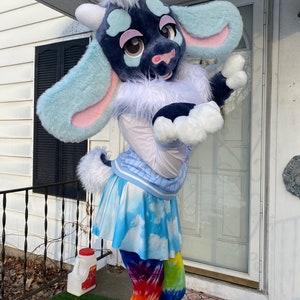 Puede incluir: Un juguete de peluche de cabra azul y blanco que lleva una camisa blanca con un patr&oacute;n de rayas azul y blanco, una falda azul y blanco con estampado de nubes y leggings de color tie-dye arco&iacute;ris.