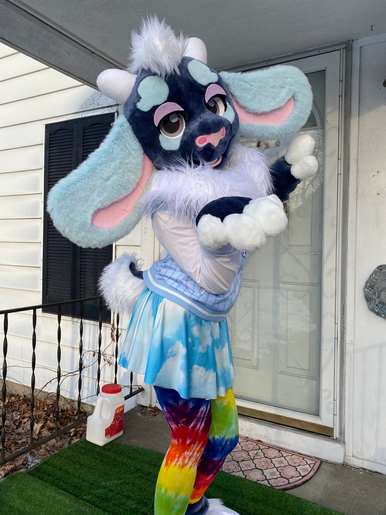 Puede incluir: Un juguete de peluche de cabra azul y blanco que lleva una camisa blanca con un patr&oacute;n de rayas azul y blanco, una falda azul y blanco con estampado de nubes y leggings de color tie-dye arco&iacute;ris.