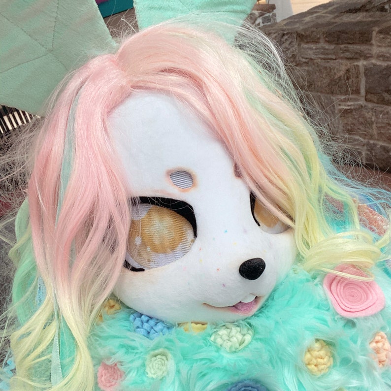 Puede incluir: Un juguete de peluche con una cara blanca, pelo rosa y amarillo y un cuerpo peludo azul claro y verde. El juguete tiene ojos grandes y oscuros y una peque&ntilde;a nariz negra.