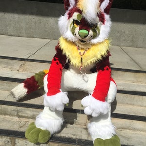 Puede incluir: Un disfraz de lobo peludo rojo, blanco y verde con una melena amarilla y patas verdes. El lobo est&aacute; sentado en un conjunto de escaleras.