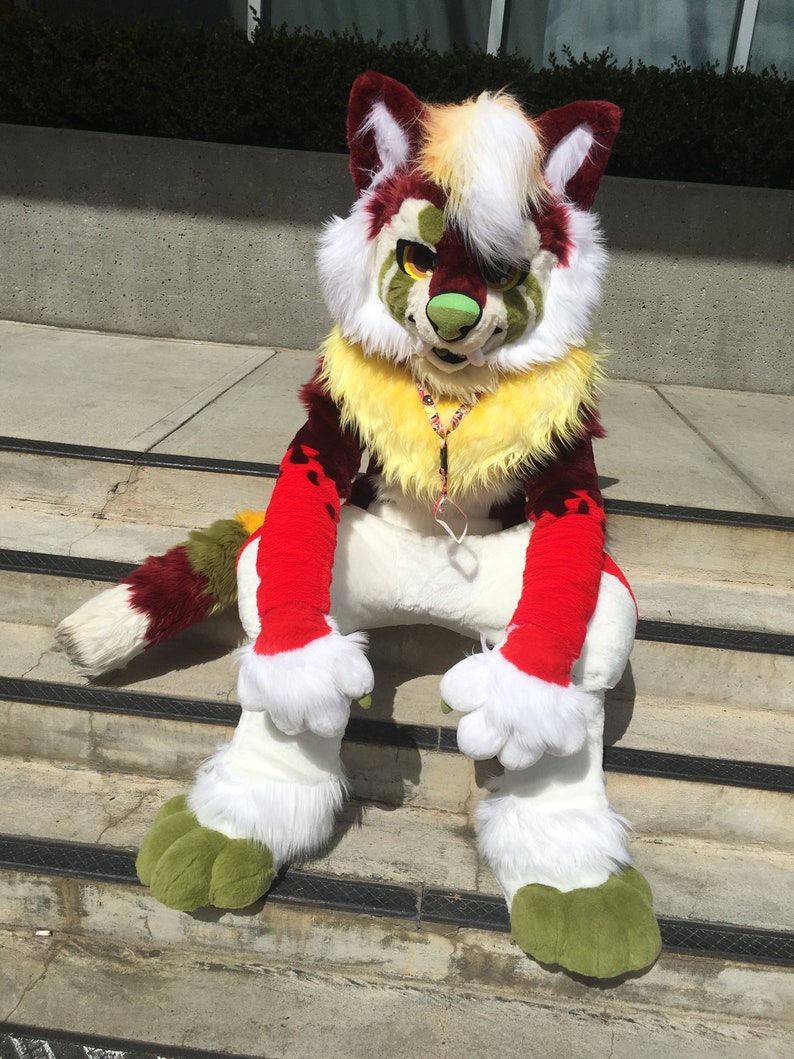 Puede incluir: Un disfraz de lobo peludo rojo, blanco y verde con una melena amarilla y patas verdes. El lobo est&aacute; sentado en un conjunto de escaleras.