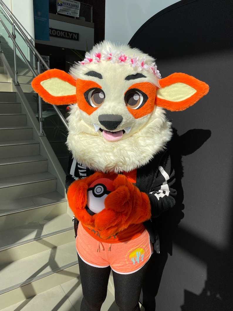 Puede incluir: Un disfraz de peluche blanco y naranja con la lengua rosa fuera. El disfraz lleva una chaqueta negra con rayas blancas y pantalones cortos naranjas con un dise&ntilde;o de puesta de sol y cactus. El disfraz sostiene una Pok&eacute; Ball roja y blanca.