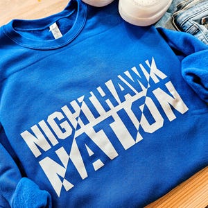 May include: A royal blue sweatshirt with the words "Nighthawk Nation" printed in white on the front. The text is in a bold, blocky font and is angled diagonally across the sweatshirt.