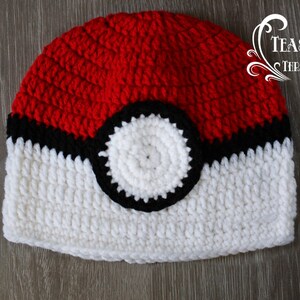 May include: A handmade crochet beanie in the style of a Poké Ball. The hat features a red top, a black stripe, a white bottom, and a white circle with a black outline. The hat is designed to resemble a popular animated character.