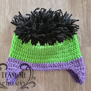 May include: A handmade crochet hat in green, purple, and black, resembling a character's hair. The hat features a green crown, a purple band, and black yarn strands at the top. The hat has ear flaps and is designed for a child.
