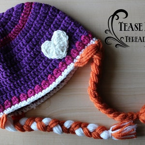 May include: A handmade crochet hat in purple, with a white heart applique and orange braided ties. The hat features pink and white trim and the text "TEASE ME THREADS".