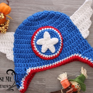 May include: A handmade blue crochet hat with a white star emblem and red and white trim. The hat features white crochet wings on each side. The hat is designed to resemble a superhero's helmet. The hat is a handmade item.