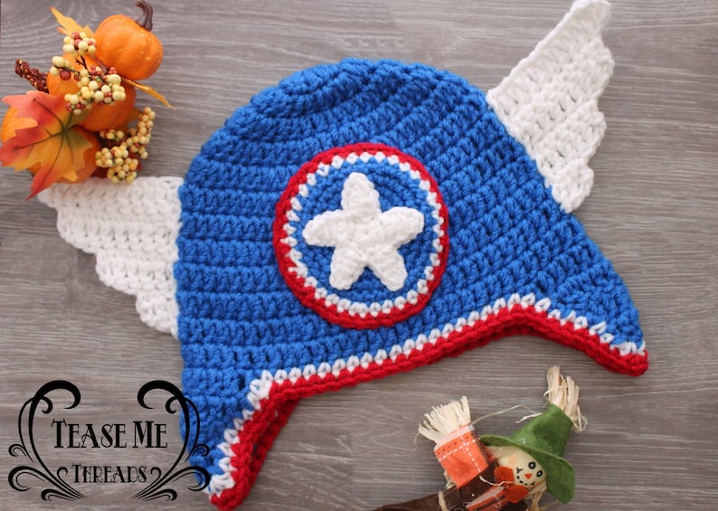 May include: A handmade blue crochet hat with a white star emblem and red and white trim. The hat features white crochet wings on each side. The hat is designed to resemble a superhero's helmet. The hat is a handmade item.