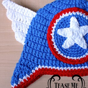 May include: A handmade crochet hat in blue, white, and red, featuring a shield design with a white star and a white wing. The hat is likely for a baby or child. The text "TEASE ME THREADS" is visible.