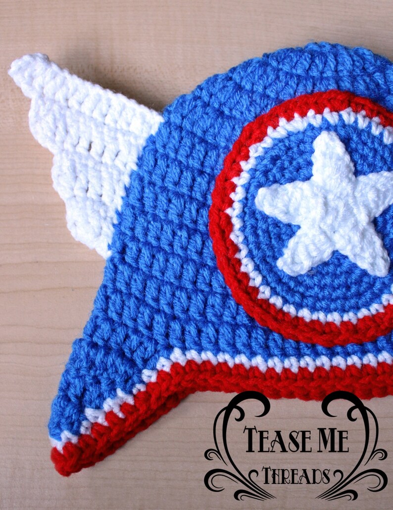 May include: A handmade crochet hat in blue, white, and red, featuring a shield design with a white star and a white wing. The hat is likely for a baby or child. The text "TEASE ME THREADS" is visible.