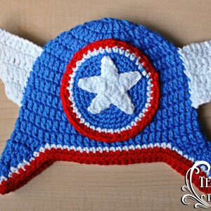 May include: A handmade blue, red, and white crochet hat with a star and shield design. The hat features white crochet wings on each side. The hat is a superhero-themed accessory.