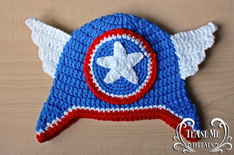 May include: A handmade blue, red, and white crochet hat with a star and shield design. The hat features white crochet wings on each side. The hat is a superhero-themed accessory.