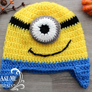 May include: A yellow, blue, and black crocheted hat resembling a Minion character. The hat features a large, single eye, a smiling mouth, and a blue band at the bottom. The hat is handmade and suitable for children.