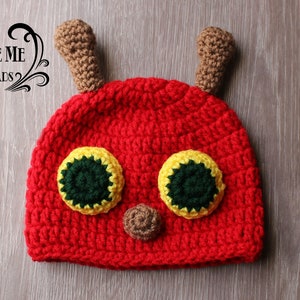 May include: A handmade red crochet hat with a caterpillar design. The hat features large yellow and green eyes, a brown nose, and two brown antennae. The hat is designed to be worn by a baby or toddler.