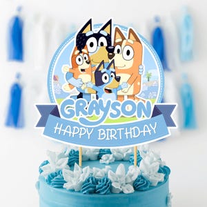 Op de afbeelding: Een ronde taarttopper met de Bluey-tekenfilmfiguren in blauw, oranje en crème. De topper bevat de naam Grayson en de tekst Happy Birthday op een blauwe banner, ondersteund door twee houten stokjes op een blauwe taart.
