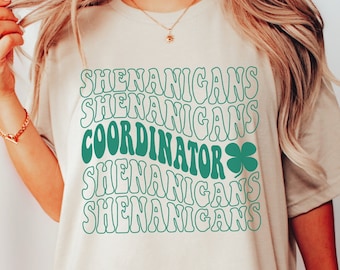 May include: A beige t-shirt with the words "SHENANIGANS COORDINATOR" in a wavy, retro font, with a green shamrock graphic. The shirt is a relaxed fit, perfect for St. Patrick's Day or casual wear.