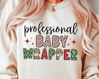 May include: Cream-colored sweatshirt with the text "professional BABY WRAPPER" in a festive font. The word "BABY" is in red and white stripes, while "WRAPPER" is in green and red. A fun holiday-themed top.
