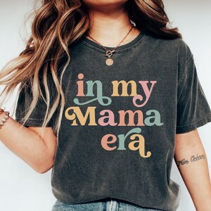 Op de afbeelding: Een donkergrijs T-shirt met de tekst "in my mama era" in kleurrijk retro-lettertype. Het shirt wordt gedragen door een persoon met lang bruin haar en wordt gecombineerd met een blauwe jeans en witte sneakers. De tekst "size S PEPPER" is zichtbaar onderaan de afbeelding.