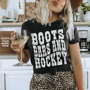 May include: A woman wearing a black t-shirt with the text "Boots Bars and Hockey" in white. She is wearing a brown hat and leopard print pants. She is holding a glass of white wine.