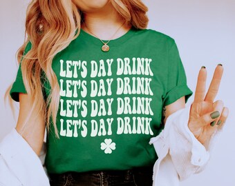 Może przedstawiać: Kobieta w zielonej koszulce z napisem "Let's Day Drink" powtórzonym cztery razy. Ma na sobie czarne dżinsy i białe trampki. Siedzi na czerwonym stołku ze skrzyżowanymi nogami.