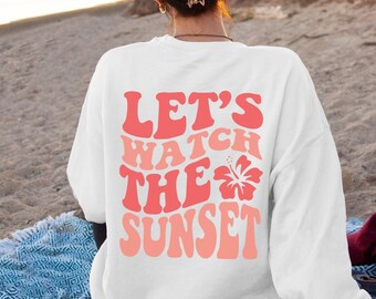 May include: White sweatshirt with the text "LET'S WATCH THE SUNSET" in shades of red and coral, with a hibiscus flower graphic. The sweatshirt is worn on a beach, with a blue patterned blanket and a camera visible.