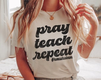 May include: A white t-shirt with the text "pray teach repeat" in black, retro-style font. The text is centered on the shirt and includes the hashtag "#homeschoolmom" below it.