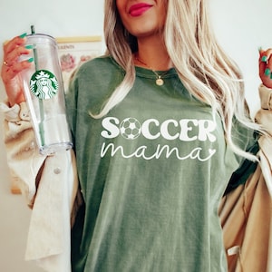 May include: A moss green oversized t-shirt with the text "soccer mama" in white with a white soccer ball graphic. The shirt is shown on a model wearing a cream colored jacket.