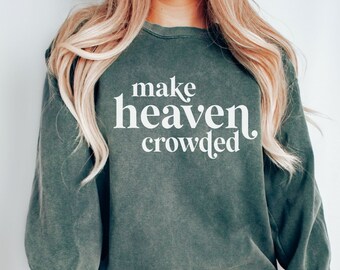 May include: Blue spruce-colored sweatshirt with the text "make heaven crowded" in white. The long-sleeved top is worn with gray shorts and black high-top sneakers. The sweatshirt is a relaxed fit.