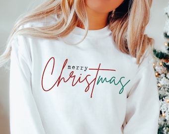 Op de afbeelding: Witte crewneck sweatshirt met de woorden "merry Christmas" in een scriptlettertype. Het woord "merry" is donkerrood en "Christmas" is groen. Een feestelijk kledingstuk voor de feestdagen.