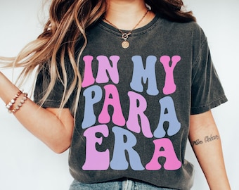 May include: A dark gray t-shirt with the text "IN MY PARA ERA" in pink and blue letters. The shirt is being worn by a person with long brown hair. The text is in a retro font style.