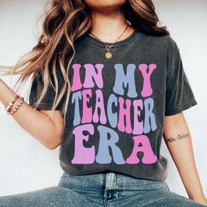 May include: A dark grey t-shirt with the text "In My Teacher Era" in pink and blue letters. The shirt is being worn by a person with long brown hair. The text "size S PEPPER" is visible at the bottom of the image.