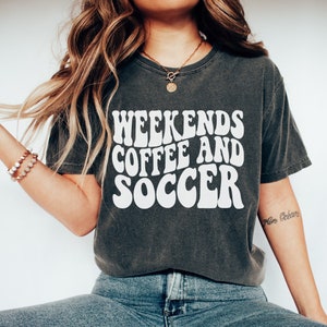 May include: A charcoal grey t-shirt with a white graphic that reads "Weekends Coffee and Soccer" in a retro font. The shirt is being worn by a person wearing blue jeans and white sneakers. The text "Size S" is visible in the bottom right corner of the image.