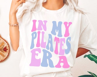 May include: White t-shirt with the text "IN MY PILATES ERA" in pink and light blue retro font. The shirt is a casual style, suitable for everyday wear. The image also shows white shorts and decorative items.