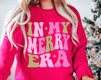 May include: A pink sweatshirt with the text "IN MY MERRY ERA" in green and pink letters. The letters have a retro, 70s style font.