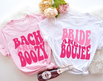 Op de afbeelding: Twee roze en witte T-shirts met de tekst "Bach and Boozy" en "Bride and Boujee" in een retro-lettertype. De shirts liggen op een wit, pluche oppervlak met een fles mousserende wijn.