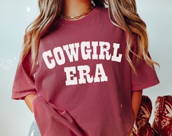 May include: A maroon t-shirt with the text "COWGIRL ERA" in white. The shirt is oversized and has a relaxed fit.